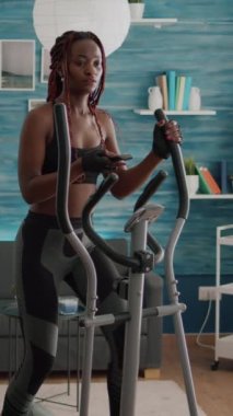 Verical video: Spor kıyafetlerinde siyah derili bisikletçi kadın oturma odasında vücut ağırlığı sırasında eliptik bisiklet kullanarak vücut kaslarını çalıştırıyor. Atletik formda yetişkin sağlıklı olmanın tadını çıkarıyor.