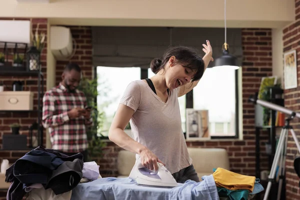 Woman singing loudly effusively while doing house chores, african american husband laughs amused in background listening. Housewife happily doing housework, culturally diverse couple.