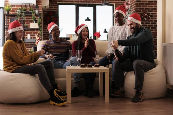 Cheerful colleagues drinking wine at christmas party, pouring alcohol in glasses to celebrate winter holiday festivity in workplace with xmas decorations and festive ornaments.