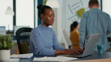 Portrait of african american woman typing using laptop keyboard and smiling at camera sitting at desk in busy startup office. Casual business employee working relaxed with sales statistics.