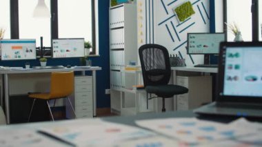 Empty startup office with multiple computer screens displaying financial statistics and closeup on laptop with moving charts on desk. Selective focus on portable computer with business erp software.
