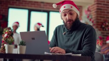Genç yetişkin beyin fırtınası fikirleri dizüstü bilgisayarda rapor yaratıyor, Noel Baba şapkası takıyor ve noel ağacı takıyor. Bayram döneminde bilgisayar üzerinde araştırma verileriyle çalışan şenlikli bir adam. El kamerasıyla..