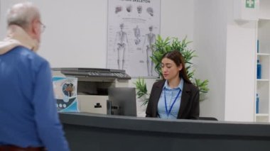 Receptionist and doctor helping old patient with cervical neck collar at hospital reception desk, wearing medical foam after injury. Diverse team giving support to injured senior man. Tripod shot.