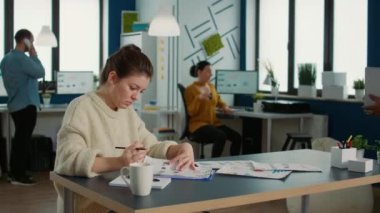 African american woman holding laptop asking colleague looking at clipboard with charts for advice regarding financial data in busy office. Two colleagues analzying market share brainstorming ideas.