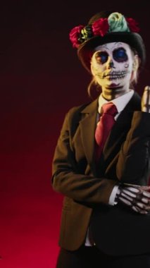 Dikey video: Cavalera Catrina takım elbiseli ve şapkalı gizemli görünümlü festival kafatası vücut sanatı giymiş, Santa Muerte Dios de los Muertos 'u stüdyoda kutluyor. Kutsal Meksika varlığı gibi görünen bir kadın..