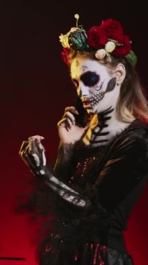 Dikey video: Santa Muerte modeli akıllı telefondan konuşuyor, cadılar bayramı makyajı yapıyor ve stüdyoda çiçek tacını takıyor. La Cavalera Catrina akıllı telefonu kullanarak Dios de los muertos Mexico 'da sohbet ediyor.