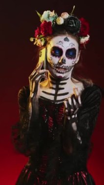 Dikey video: Genç korkutucu kadın akıllı telefondan konuşuyor, cadılar bayramı makyajı yapıyor ve stüdyoda çiçek tacını takıyor. La Cavalera Catrina, Dios de los Muertos Meksika tatili hakkında konuşmak için akıllı telefonu kullanıyor.