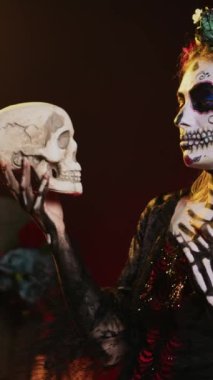 Dikey video: Korku Noel Baba stüdyoda kafatasına bakıyor, saçbantlı ürkütücü cadılar bayramı kostümü giyiyor. Dios de los Muertos 'ta siyah beyaz makyajlı kutsal bir varlık gibi davranıyorlar.