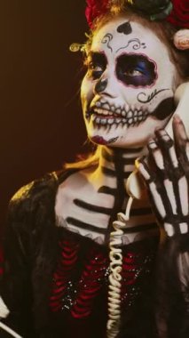 Dikey video: Ölüm Tanrıçası stüdyoda sabit telefon ile konuşuyor, telefon kablosuyla konuşuyor ve kafatası makyajı yapıyor. Santa Muerte kostümlü kadın kutsal Meksika ritüelini kutluyor.