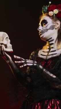 Dikey video: Stüdyoda kafatasıyla konuşan kutsal tanrıça, Meksika cadılar bayramı geleneğini kutlamak için korkutucu ve dehşet verici davranıyor. Korkunç festival kostümü içinde vücut sanatı olan bir Noel Baba.