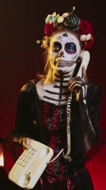Dikey video: Ürkütücü kadın sabit hatlı telefona kabloyla cevap veriyor, kafatası makyajı ve vücut sanatı yaparken ofis telefonuyla konuşuyor. Dios de los muertos 'ta Noel Baba' yı canlandıran kadın.