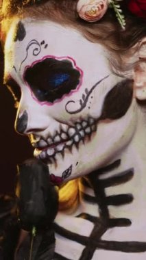 Dikey video: Ölüm kostümü ve çiçek çelengi giyen göz kamaştırıcı tanrıça stüdyo arka planında, kutsal Noel Baba Muerte gibi davranıyor. La Cavalera Catrina Kafatası vücut sanatı, Meksika kültürü ile ölü kadın