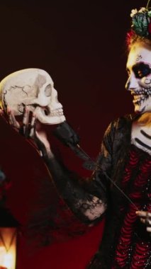 Dikey video: Kafatası ve siyah güllerle flört eden korkunç kadın model, Meksika tatilini kutlamak için geleneksel vücut sanatı giyiyor. Dios de los Muertos ayinindeki ölüm tanrıçasına benziyor.