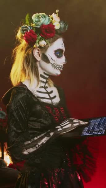 垂直ビデオ ラップトップを保持しているハロウィーンの衣装を着た気味の悪い女性 スタジオで伝統的なメキシコの休日にポーズ 頭蓋骨を身に着けていることは死者の日に体の芸術を構成し ワイヤレスPcを使用して閲覧する — ストック動画