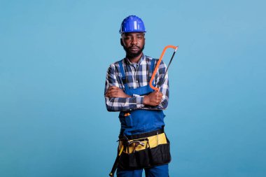 Ciddi inşaat işçisi testereyle mavi arka planda izole edilmiş kameraya bakıyor. Marangoz iş aletlerini üniforma ve koruyucu miğfer giyerek tutuyor..