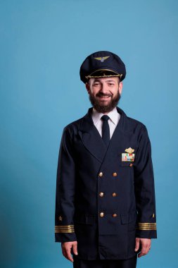Havacılık üniforması ve şapka giyen bir kaptan portresi, kameraya bakan bir uçak pilotu. Profesyonel ceketli havacı, stüdyoda mavi arka planda uçuş ekibi üyesi.