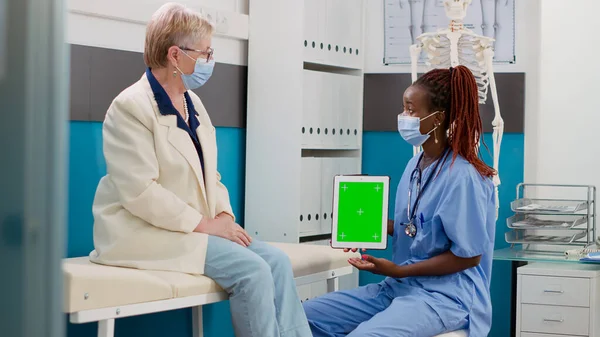 Nurse and patient with face mask looking at greenscreen on digital tablet, doing consultation in office. Assistant holding display with isolated chromakey template and blank copyspace.