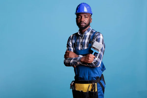 Serious professional painter holding brush wearing overalls and protective helmet, arms crossed over chest. African american construction worker against blue background in studio shot.