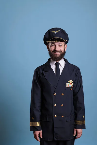 Portrait of airplane captain wearing aviation uniform and hat, plane pilot looking at camera. Aviator with badge on professional jacket, aircrew member on blue background in studio