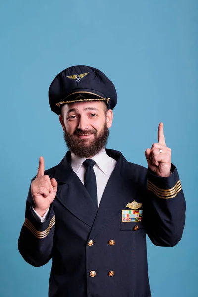 Portrait of smiling aviator pointing up with index fingers, advertising product, wearing aviation uniform. Plane pilot looking at camera with forefingers showing upwards, promotion gesture