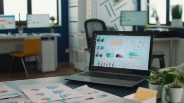 Closeup of laptop screen showing moving charts and financial data in empty modern office in startup company. Interior of marketing department with portable computer showing business data on desk.