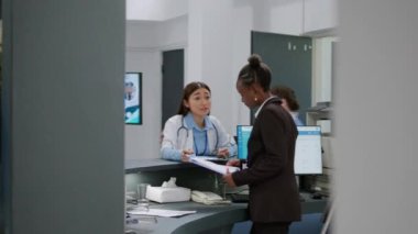 Hastane resepsiyonundaki sağlık raporları üzerinde çalışan çeşitli tıbbi çalışanlar. Asyalı doktor ve Afrikalı Amerikalı resepsiyonist kayıt masasında..