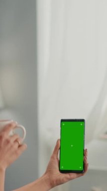 Dikey video: Ofis masasında kahve içen serbest çalışan kadın meslektaşıyla konuşuyor sahte yeşil ekran krom anahtar akıllı telefon kullanıyor. Çevrimiçi bilgi arayan beyaz kadın izole telefon kullanıyor
