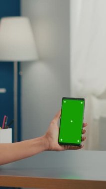 Dikey video: Sosyal medya kadını dokunmatik ekranlı telefonu modern ofis masasında oturan yeşil ekran krom anahtarıyla tutuyor. Çevrimiçi uygulamalarda çalışan beyaz kadın izole edilmiş akıllı telefon kullanıyor.