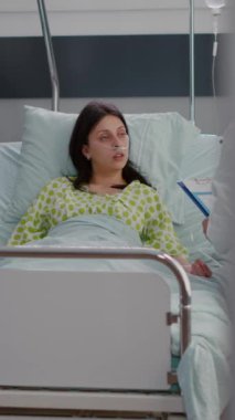 Dikey video: hastane koğuşunda yatağında nazal oksijen tüplü beyaz kadın. Pratisyen doktor hastayla hastalık testi sonuçlarını panoya yazma konusunda tartışıyor.