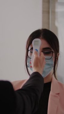 Dikey video, kadının vücut ısısını ölçerken tıbbî termometreyle ölçüp kovid 19 'u önlemek için. İş yerinde, Coronavirus sırasında sosyal mesafeyi koruyan iş arkadaşları koruma maskesi takıyor.