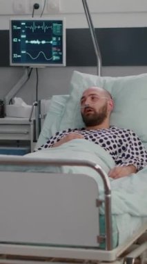 Dikey video: Hasta adam yatakta oksijen tüpüyle oturuyor ve panoya hastalık tedavisi yazarken pratisyen doktora hastalık belirtilerini açıklıyor. Tıp hemşiresi oksimetreyi inceliyor.