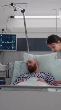 Dikey video: Afro-Amerikan doktor hasta adam hastalıkları tedavisi ile ilgili belirtileri panoya yazma konusunda uzman doktorla tartışıyor. Hemşire hasta yatağı ayarlayıp hastanede çalışıyor.
