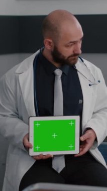 Dikey video: Beyaz önlüklü uzman hasta kadınla hastalık tedavisi hakkında konuşuyor. Elinde yeşil ekran kroma anahtar tableti bilgisayarı ile izole ekran tutan bir uygulayıcı