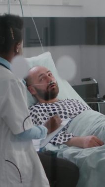 Dikey video: Afro-Amerikalı doktor hastane koğuşundaki iyileşme randevusu sırasında hastalık belirtilerini izleyen hasta adamı kontrol ediyor. Doktor kalp atışlarını analiz ederken hasta yatakta oturuyor.