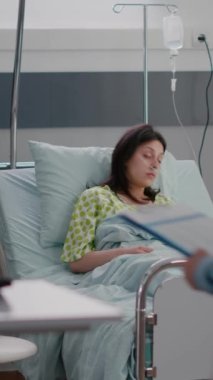 Dikey video: Pratisyen hekim hastane koğuşundaki hastalık randevusu sırasında yatağında dinlenen hasta kadını kontrol ediyor. Doktor, tıbbi oksimetre izleme nabız ölçümü yapıyor.
