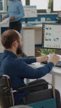 Video dikey: Felç olmuş, hareketsiz, felçli iş adamı finansal rapor yazıyor ve modern girişimci iş yerinde tekerlekli sandalyede oturan iş arkadaşıyla konuşuyor