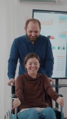 Video dikey: Özürlü sakat iş arkadaşı ile tekerlekli sandalyeyi iten neşeli genç adam yeni açılan finans ofisinde gülümserken. İş arkadaşları iş yerinde gülüp eğleniyor.