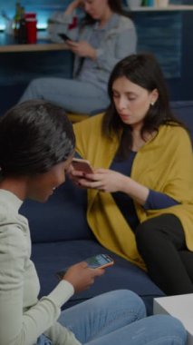 Dikey video: Karışık ırk arkadaşları kanepe holdinglerinde el ele oturuyor internette geziniyor, rahatlama partisi sırasında çevrimiçi video izliyorlar. Çok ırklı gruplar birlikte vakit geçiriyorlar.