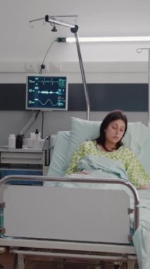 Dikey video: Hastane koğuşundaki rehabilitasyon sırasında hasta kadını izleyen kadın doktor, oksimetrenin hastalık uzmanlığını kontrol ettiğini gösteriyor. Hasta yatağında dinleniyor, hastalık tedavisini bekliyor.