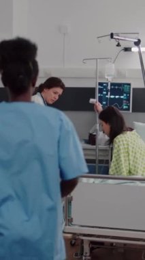 Dikey video: Hastanın hayati değerlerini kontrol eden tıbbi ekip kalp atış hızını kontrol ediyor. Hastanenin koğuşunda sıvı akışı için serum torbasına yardım ediyor. Klinik sırasındaki hasta kadın sağlık hizmetlerini bekliyor.