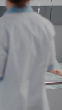 Dikey video: Doktor, hastane koğuşundaki kardiyoloji randevusu sırasında hasta kadını izleyerek tıbbi oksimetrenin nabız uzmanlığını kontrol etti. Hasta yatakta dinleniyor, bekliyor.