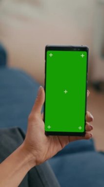 Dikey video: Kafkas kadın oturma odasında kanepede otururken internet uygulamasında sosyalleşirken yeşil ekran krom anahtar görüntüsünü taklit ederek telefon kullanıyor. Bir kadın izole edilmiş aygıtla ağda geziniyor