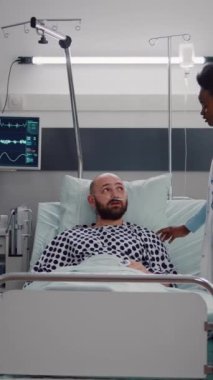 Dikey video: Hastanenin koğuşunda hasta, yatağında dinlenirken doktorlarla tartışıyor. Panoya hastalık tedavisi yazan pratisyen. Hastalık belirtilerini inceleyen.