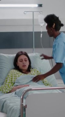 Dikey video: Siyahi tenli asistan, hastane koğuşunda çalışırken hasta kadın yatağı düzenliyor, pratisyen doktor hastalık belirtilerini kontrol ediyor