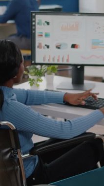 Dikey video: İşlek odaklı Afrikalı felçli kadın yönetici bilgisayardan grafikleri ve finansal istatistikleri analiz ediyor, işe başlama ofisinde tekerlekli sandalyede oturarak yazıyor