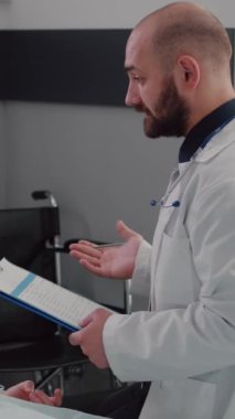 Dikey video: Hastane koğuşunda çalışan pratisyen hekim, pano izleme hastalıkları teşhisi konusunda uzmanlık yazısı yazıyor. Hasta hasta yatağında dinleniyor ve hastalık tedavisi hakkında konuşuyor.