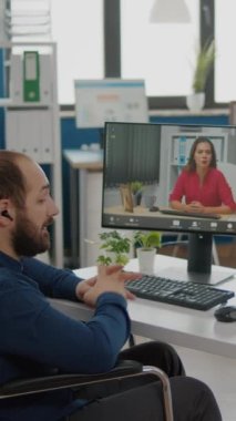 Video dikey: Tekerlekli sandalyede oturan iş ortağıyla ofis planlamasında video konferansı sırasında iş arkadaşıyla konuşan engelli, felçli yönetici