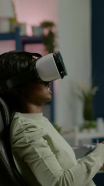 Dikey video: Güçlü bilgisayarda video oyunu oynayan siyah kadın oyuncu VR kulaklık takıyor. Heyecanlı oyuncu evdeki sanal turnuva oyunları için kablosuz denetleyici kullanıyor