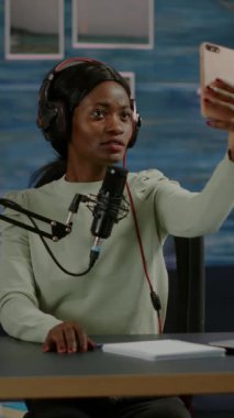Dikey video: Akıllı telefon kullanan Afrikalı kadın eğlence sektörü kayıt bölümünde selfie çekiyor. Çevrimiçi prodüksiyon internet podcast 'i sunucunun canlı yayın yaptığını gösteriyor, dijital kayıt