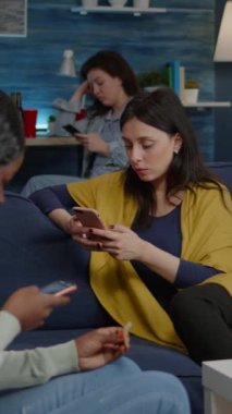Dikey video: Sürpriz parti sırasında mobil hız yapma süresini kullanarak internette sörf yaparken koltukta dinlenen çok ırklı arkadaşlar. Çevrimiçi komik film izleyen çok ırklı bir grup insan.